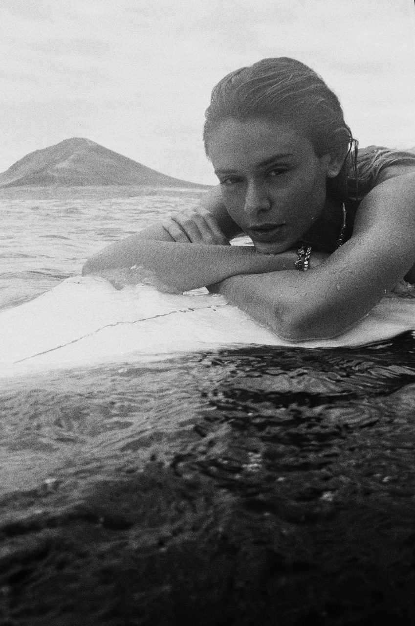 Sergio Felipe García. Black and white portrait of a woman lay down on a surfboard in the sea.