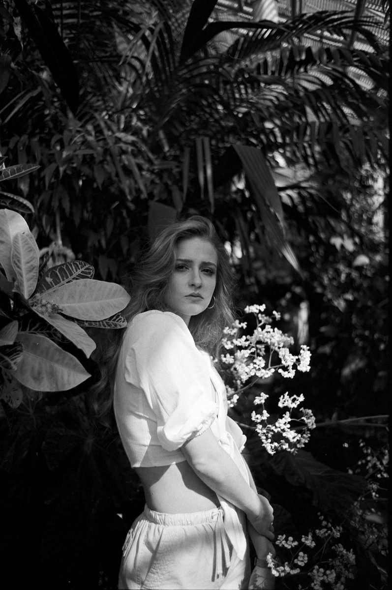 Michèl Passin. Black and white portrait of a woman stood in front of many plants.