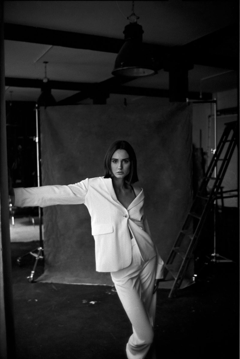 Michèl Passin. Black and white portrait of a woman stood up in front of a backdrop leaning against the wall.