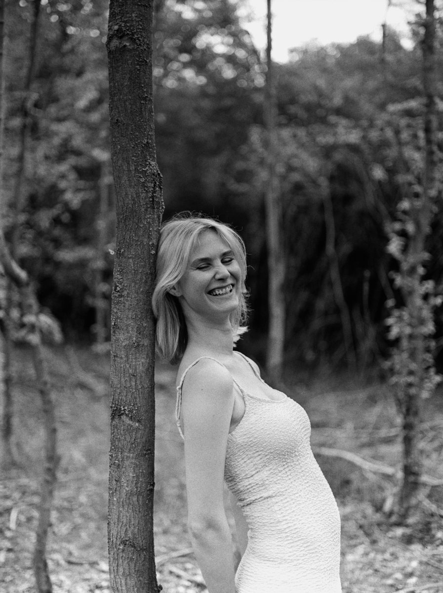 Michèl Passin. Black and white portrait of a woman in a white dress smiling and leaning against a tree.