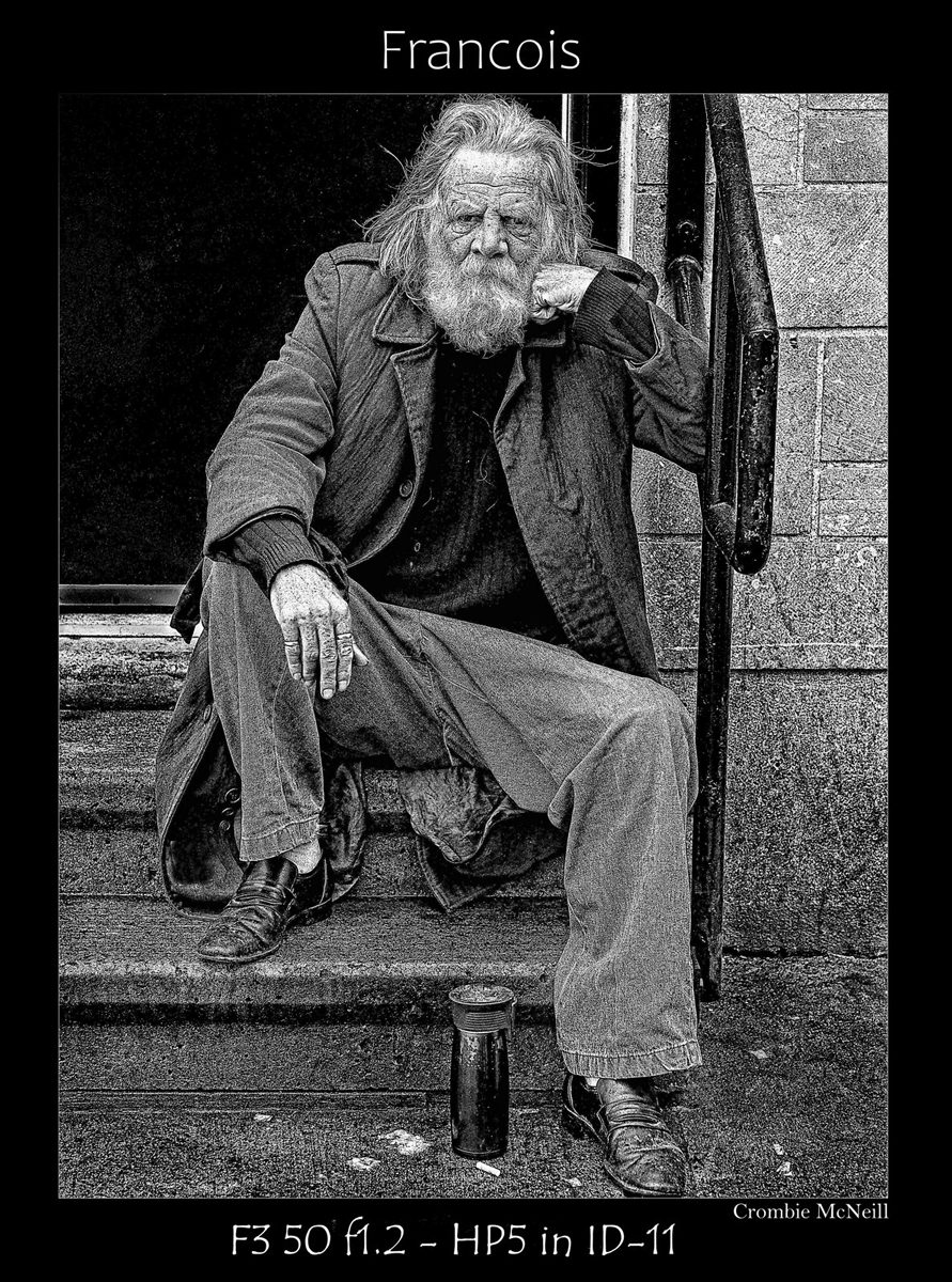 Crombie McNeill. Black and white shot of an old man with a beard sat on a step leaning against the banister outside.