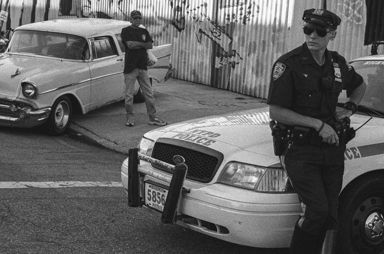 Larry Niehues. Black and white shot of a police officer leaned on a police car.