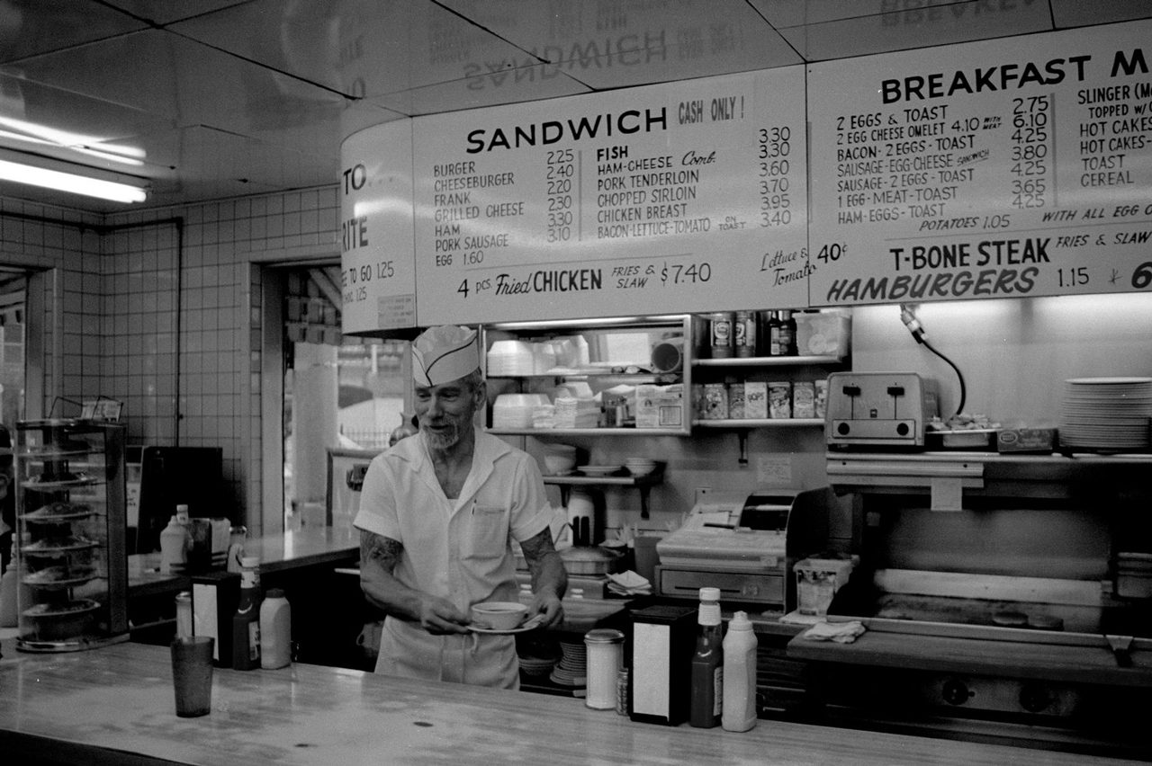 Larry Niehues. Black and white documentary shot of a chef behind the counter in a cafe.