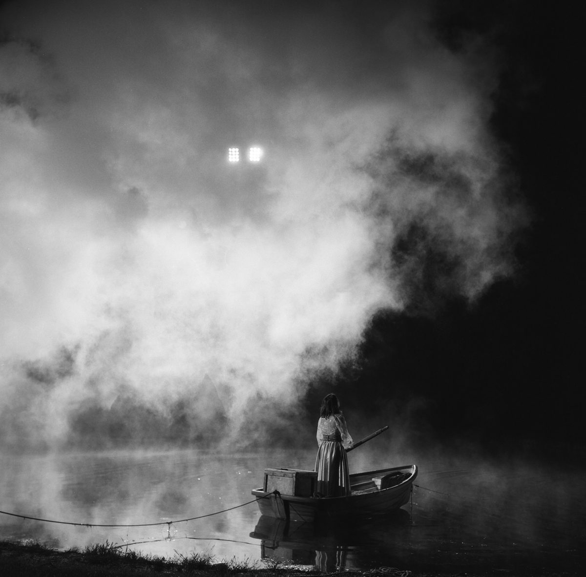 Hannah Baker. Black and white image of a woman on stood on a boat going down a river with misty clouds.