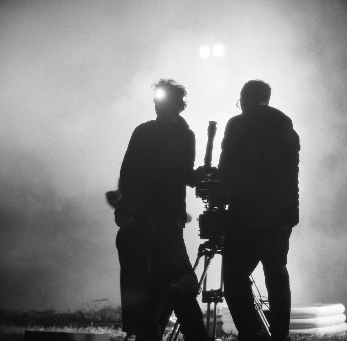 Hannah Baker. Black and white silhouette of two men on set with white smoke surrounding them holding filming equipment.