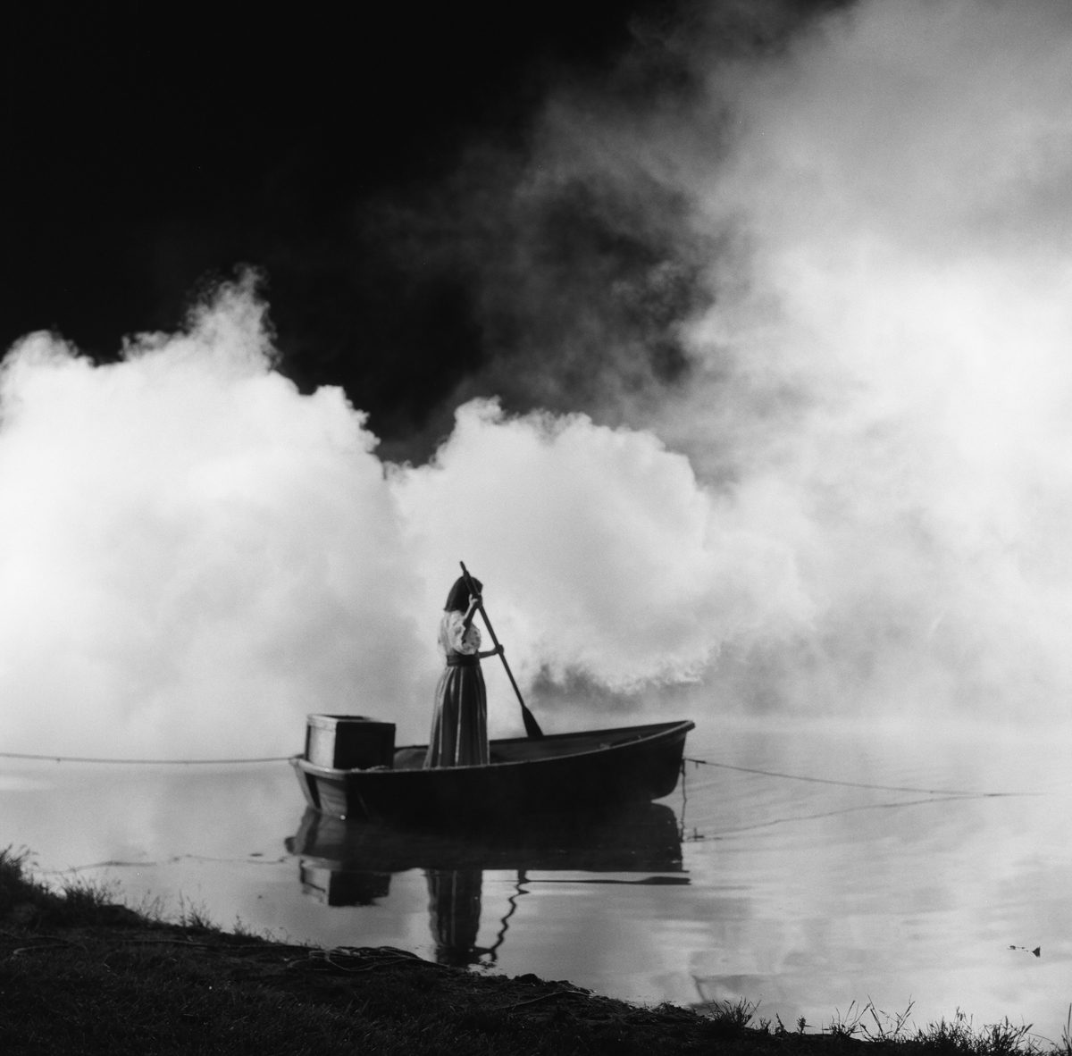 Hannah Baker. Black and white image of a woman on stood on a boat going down a river with misty clouds.