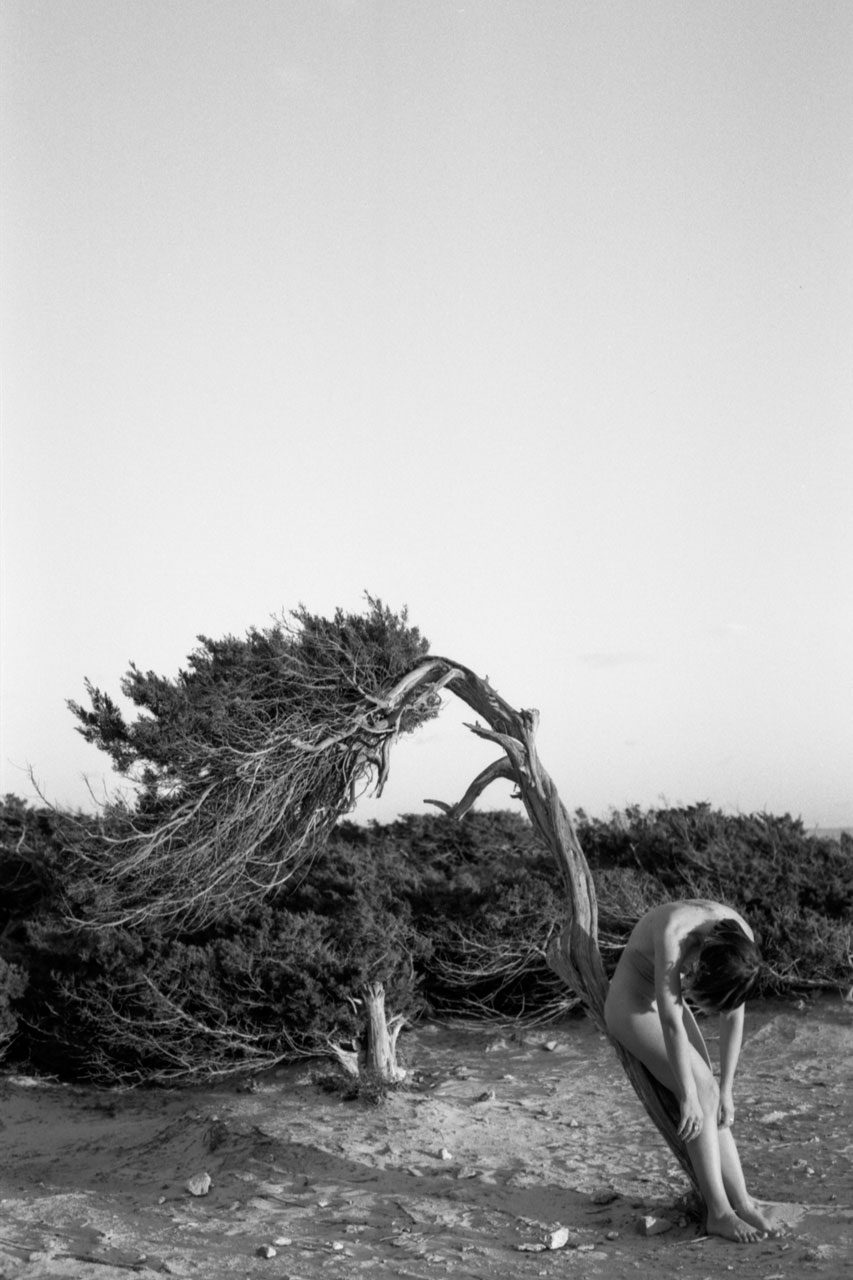 Clarissa Sofia. Black and white image of a naked woman bent over in front of a bent tree.