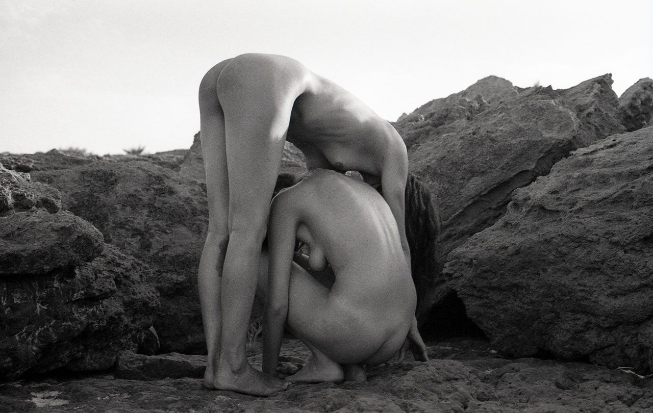 Clarissa Sofia. Black and white image of two women naked in front of a rock bent over each other